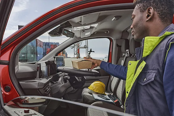 Fahrerhaus des roten Ford Transit Custom, sichtbar durch geöffnete Fahrertür, Mann in Warnweste stellt Lunchpaket auf das Lenkrad.