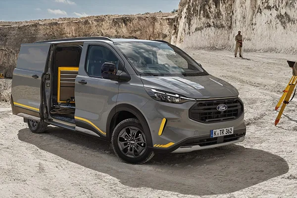 Grauer Ford Transit Custom parkt mit geöffneter Seitentür in einem Steinbuch mit Person und Messgerät im Hintergrund.