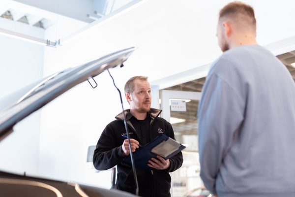 Serviceberater mit Klemmbrett in der Hand steht gemeinsam mit Kunden vor der geöffneten Motorhaube des Kundenfahrzeugs.
