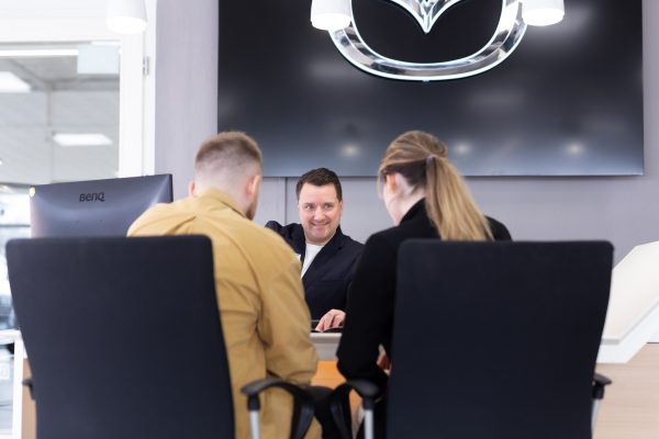 Mazda-Verkäufer sitzt am Tisch mit zwei Kunden und lächelt sie freundlich an.
